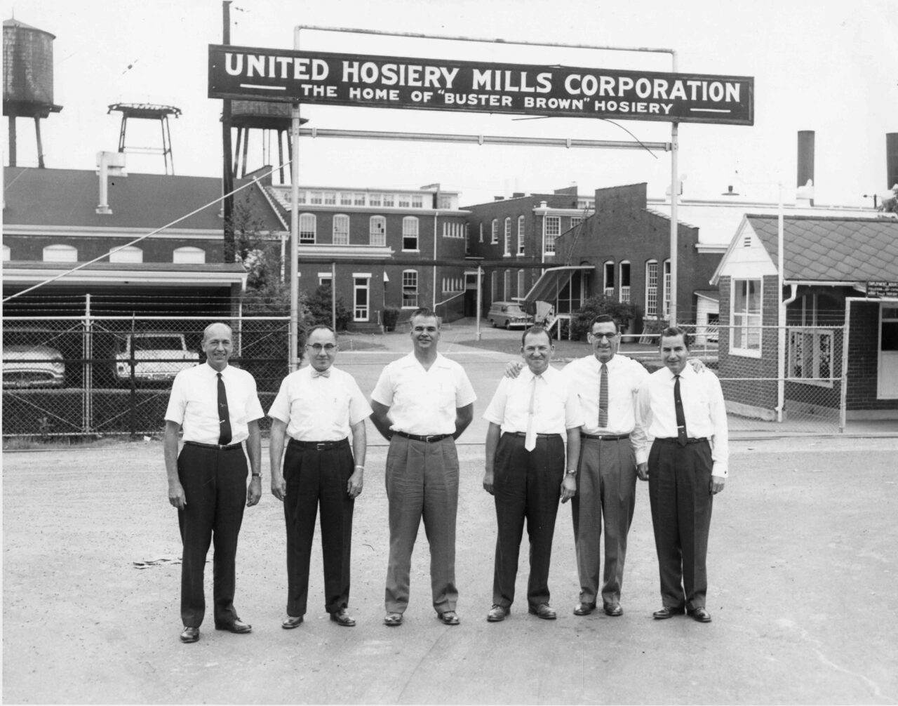 Black-and-white photo of six men standing in front of the United Hosiery Mills Corporation entrance sign reading “The Home of ‘Buster Brown’ Hosiery.” The men, dressed in white shirts and slacks, pose side by side, some with arms around each other, smiling at the camera. Behind them are factory buildings, parked cars, a chain-link fence, and water towers.