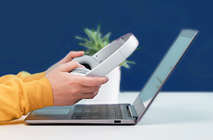 Side view of slim laptop and hands with wireless headphones on grey desk. Blue background.